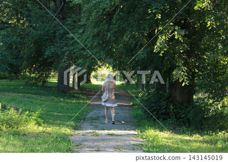 Blonde Woman in Light Summer Dress and Bag Walking Down Sun-Dappled Park Alley, Seen From Behind Blonde Woman in Light Summer Dress and Bag Walking Down Sun-Dappled Park Alley, Seen From Behind 143145019