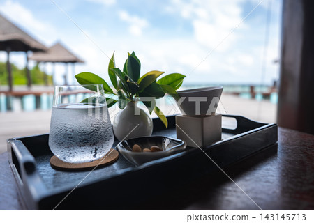 A refreshment tray with water, nuts, teacup, and vase sits on a dark table by a lagoon in Maldives, with thatched roof bungalows and turquoise water softly blurred. A refreshment tray with water, nuts, teacup, and vase sits on a dark table by a lagoon in Maldives, with thatched roof bungalows and turquoise water softly blurred. 143145713