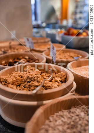 Wooden bowls hold granola, cereals, nuts, and seeds in Oberlech, Austria. Small spoons and labels sit on each bowl. Soft light and shallow depth of field focus on granola. 143145905