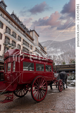 A red Mont Cervin Palace horse carriage waits by the historic hotel in Zermatt, Switzerland, with snow on alpine slopes and chalet balconies under soft overcast light. 143145961