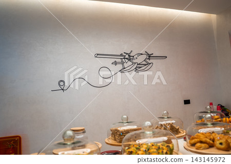 A buffet counter shows pastries and small cakes under glass cloches. A line art seaplane mural marks Maldives travel in a lounge near the TMA seaplane port in Male. A buffet counter shows pastries and small cakes under glass cloches. A line art seaplane mural marks Maldives travel in a lounge near the TMA seaplane port in Male. 143145972