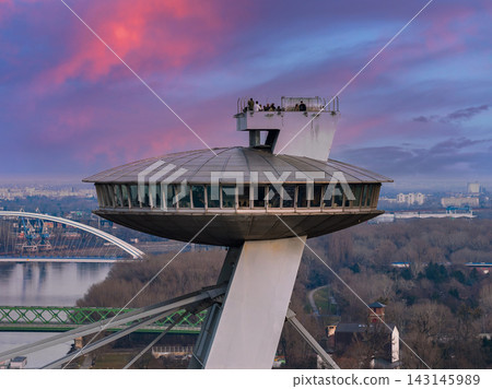 Aerial view at dusk shows the UFO Observation Tower on SNP Bridge, Bratislava, with wraparound windows and rooftop deck, above the Danube River and wooded riverbanks. Aerial view at dusk shows the UFO Observation Tower on SNP Bridge, Bratislava, with wraparound windows and rooftop deck, above the Danube River and wooded riverbanks. 143145989