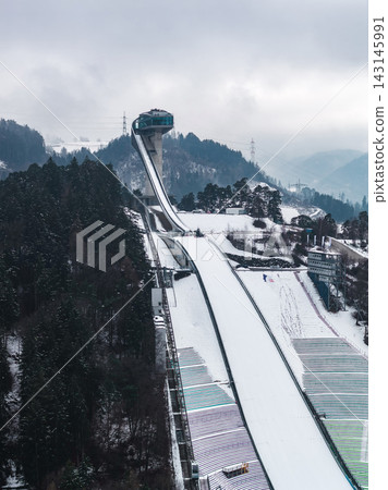 Aerial view of Bergisel Ski Jump in Innsbruck, Austria, with Zaha Hadid tower, long white inrun, stepped landing, winter snow, evergreen forests, and misty Alpine ridges. 143145991