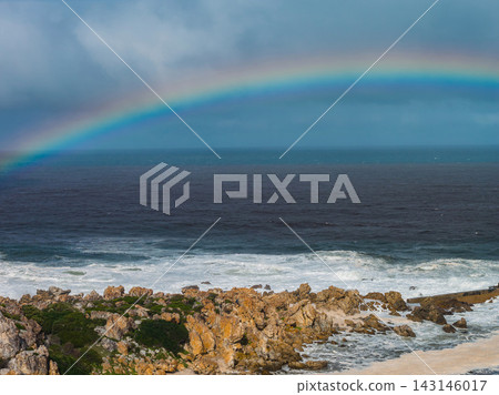 Rugged ochre rocks and coastal vegetation line the Cape Town shore as a rainbow arcs over dark choppy Atlantic water, with storm light meeting bright breaking surf. Rugged ochre rocks and coastal vegetation line the Cape Town shore as a rainbow arcs over dark choppy Atlantic water, with storm light meeting bright breaking surf. 143146017