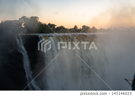Wide view of Victoria Falls at sunrise shows torrents over a basalt cliff, mist rising like smoke that thunders, treeline backlit, soft light and slight haze present. Wide view of Victoria Falls at sunrise shows torrents over a basalt cliff, mist rising like smoke that thunders, treeline backlit, soft light and slight haze present. 143146025