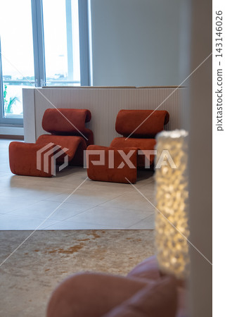 Two rust orange zigzag lounge chairs sit in a minimalist Maldives lobby with a ribbed white partition, tall windows, pale stone flooring, and a glowing textured lamp. Two rust orange zigzag lounge chairs sit in a minimalist Maldives lobby with a ribbed white partition, tall windows, pale stone flooring, and a glowing textured lamp. 143146026
