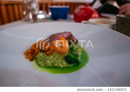 A petite seared meat and shrimp pairing rests on pea puree with orange vegetables and broccoli, on a white dish in a luxury hotel restaurant in Oberlech, Austria. A petite seared meat and shrimp pairing rests on pea puree with orange vegetables and broccoli, on a white dish in a luxury hotel restaurant in Oberlech, Austria. 143146031
