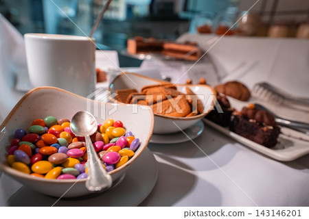 A close up dessert spread in Oberlech, Austria shows candy coated chocolates, a silver spoon, speculoos biscuits, a chocolate cake slice, and a white coffee mug under soft light. A close up dessert spread in Oberlech, Austria shows candy coated chocolates, a silver spoon, speculoos biscuits, a chocolate cake slice, and a white coffee mug under soft light. 143146201