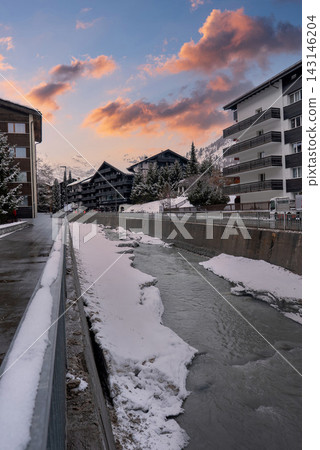 A narrow canal flows along a snowy street in Zermatt, Switzerland, with modern chalet facades, concrete embankments, misty Alps, and an overcast late winter mood. A narrow canal flows along a snowy street in Zermatt, Switzerland, with modern chalet facades, concrete embankments, misty Alps, and an overcast late winter mood. 143146204