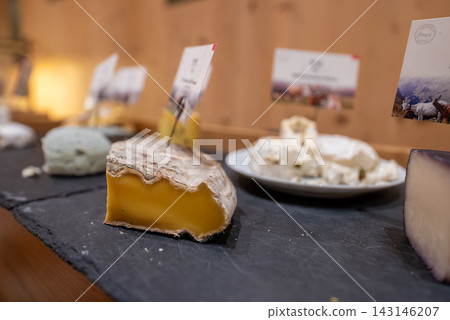 A cheese tasting display shows labeled varieties on a dark slate board. An aged orange yellow wedge with natural rind is in focus under warm indoor lighting. A cheese tasting display shows labeled varieties on a dark slate board. An aged orange yellow wedge with natural rind is in focus under warm indoor lighting. 143146207