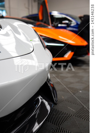 White supercar nose at left in sharp focus, orange Lamborghini Revuelto sits blurred with scissor door raised beside a blue car in an indoor detailing bay with tiles and lights. White supercar nose at left in sharp focus, orange Lamborghini Revuelto sits blurred with scissor door raised beside a blue car in an indoor detailing bay with tiles and lights. 143146238