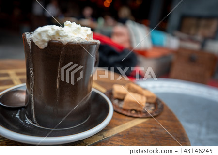 A dark ceramic mug with dripping whipped cream sits on a saucer with spoon, brown sugar cubes nearby, in a cozy lounge in Zermatt, Switzerland, daylight glow. 143146245