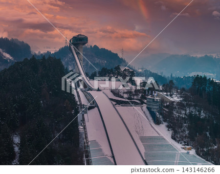 Aerial view of Bergisel Ski Jump in Innsbruck, Austria at dusk, Zaha Hadid tower above snow forests and terraced slopes, faint rainbow over misty Tyrolean mountains. 143146266