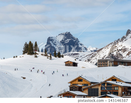 Skiers and snowboarders glide on gentle pistes by chalets and modern hotels in Lech and Oberlech, Austria, with fir trees and huts under soft winter daylight. Skiers and snowboarders glide on gentle pistes by chalets and modern hotels in Lech and Oberlech, Austria, with fir trees and huts under soft winter daylight. 143146387