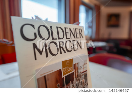 A menu titled Goldener Morgen sits on a table in Hotel Goldener Berg, Oberlech, Austria. Warm red seating, curtained windows, muted lighting, and shallow depth of field are visible. A menu titled Goldener Morgen sits on a table in Hotel Goldener Berg, Oberlech, Austria. Warm red seating, curtained windows, muted lighting, and shallow depth of field are visible. 143146467