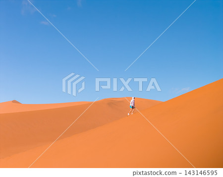 A lone hiker climbs a steep dune in Sossusvlei's Deadvlei, Namibia. Early or late light defines crisp contours and long soft shadows under a cloudless sky. A lone hiker climbs a steep dune in Sossusvlei's Deadvlei, Namibia. Early or late light defines crisp contours and long soft shadows under a cloudless sky. 143146595
