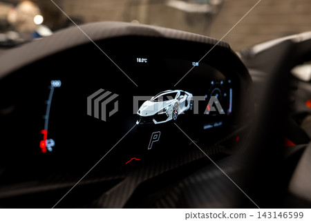 Digital instrument cluster of a Lamborghini Revuelto shows car render, gear P, 18.0 C, and tachometer gauge in a workshop with carbon fiber trim and dim light. Digital instrument cluster of a Lamborghini Revuelto shows car render, gear P, 18.0 C, and tachometer gauge in a workshop with carbon fiber trim and dim light. 143146599