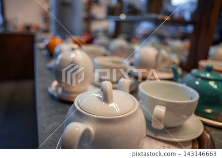 Creamy white teapots and cups sit on a counter in a hotel tea lounge in Oberlech, Austria, with a teal pot further back. Soft diffused lighting and shallow depth enhance glaze. Creamy white teapots and cups sit on a counter in a hotel tea lounge in Oberlech, Austria, with a teal pot further back. Soft diffused lighting and shallow depth enhance glaze. 143146663