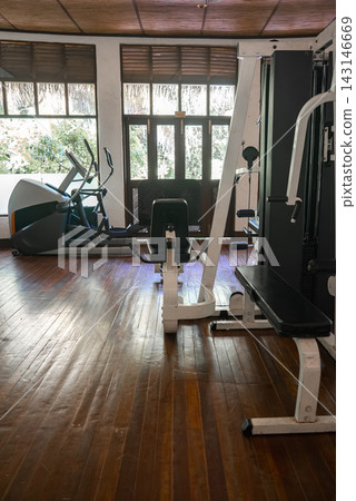 Polished wooden flooring, cable machine with bench, leg extension, and elliptical sit in soft daylight. Louvered windows and glass doors reveal tropical plants in a Maldives resort. Polished wooden flooring, cable machine with bench, leg extension, and elliptical sit in soft daylight. Louvered windows and glass doors reveal tropical plants in a Maldives resort. 143146669