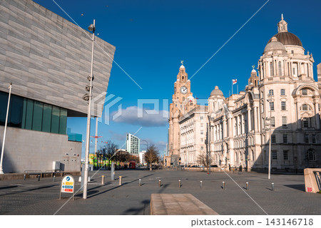 Winter sun illuminates Liverpool Pier Head. Royal Liver Building and Port of Liverpool Building stand near Museum of Liverpool. Sparse pedestrians cast long shadows. Winter sun illuminates Liverpool Pier Head. Royal Liver Building and Port of Liverpool Building stand near Museum of Liverpool. Sparse pedestrians cast long shadows. 143146718