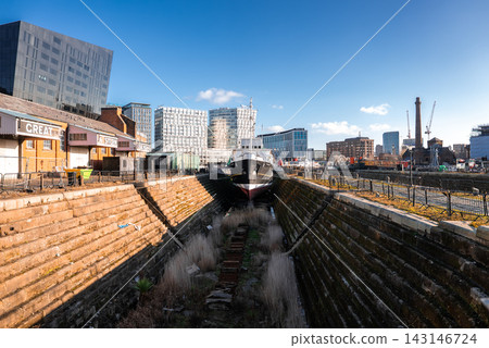 A moored ship rests in a stone dry dock at Liverpool, with Victorian warehouses, modern glass offices, the Mann Island building, a chimney, and cranes in crisp winter daylight. A moored ship rests in a stone dry dock at Liverpool, with Victorian warehouses, modern glass offices, the Mann Island building, a chimney, and cranes in crisp winter daylight. 143146724