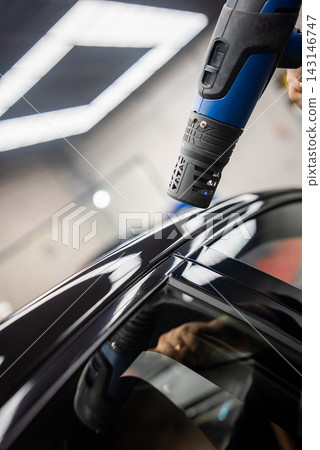 A technician uses a blue heat gun to smooth paint protection film on a glossy black luxury car roofline in a dealer workshop, with LED ceiling lights reflecting. 143146747