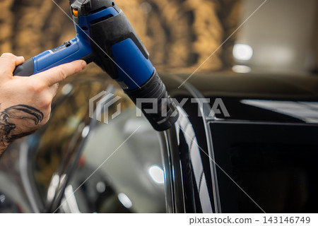 A technician uses a blue heat gun to shrink vinyl wrap on a glossy black B pillar and door frame of a luxury car inside a dealership workshop under warm indoor light. 143146749
