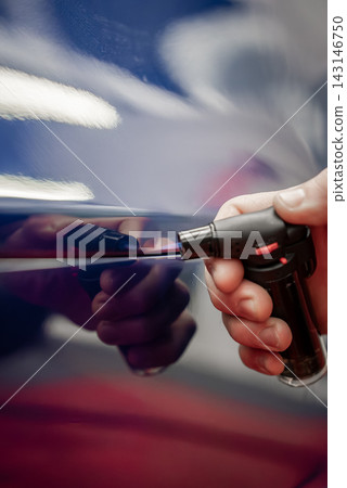 A technician uses a butane micro torch on a thin line of a glossy blue car door, with reflections, bright shop lighting, bokeh highlights, and shallow depth of field. A technician uses a butane micro torch on a thin line of a glossy blue car door, with reflections, bright shop lighting, bokeh highlights, and shallow depth of field. 143146750