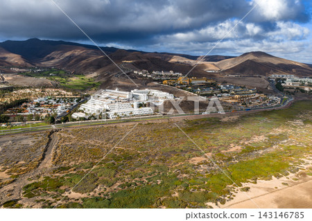 Aerial resort town near Morro Jable, Fuerteventura. Terraced hotels rise on lava slopes, golf course in valley, coastal road skirts wetland, clouds cast moving shadows. Aerial resort town near Morro Jable, Fuerteventura. Terraced hotels rise on lava slopes, golf course in valley, coastal road skirts wetland, clouds cast moving shadows. 143146785