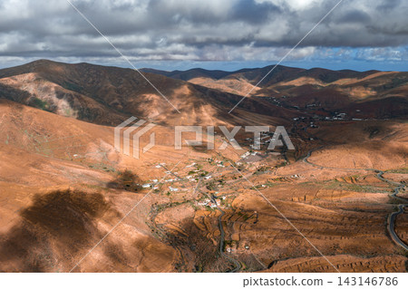Aerial view of Fuerteventura in the Canary Islands shows arid hills, winding valleys, serpentine roads, whitewashed hamlets, and low clouds over volcanic slopes. Aerial view of Fuerteventura in the Canary Islands shows arid hills, winding valleys, serpentine roads, whitewashed hamlets, and low clouds over volcanic slopes. 143146786