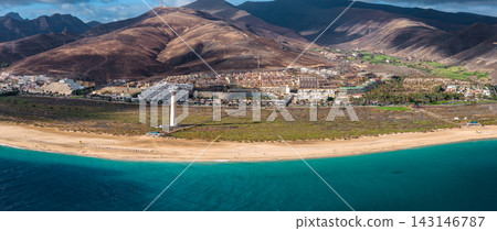 Aerial view shows Morro Jable on Fuerteventura, Jandia lighthouse, arid volcanic mountains, golden sand, turquoise water, resort buildings, and palm lined streets. Aerial view shows Morro Jable on Fuerteventura, Jandia lighthouse, arid volcanic mountains, golden sand, turquoise water, resort buildings, and palm lined streets. 143146787