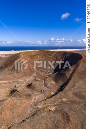 Aerial view of a rust toned caldera, badlands, and Atlantic shore in Fuerteventura. A winding dirt track crosses arid terrain under clear midday light and deep skies. Aerial view of a rust toned caldera, badlands, and Atlantic shore in Fuerteventura. A winding dirt track crosses arid terrain under clear midday light and deep skies. 143146798
