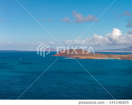 Aerial view shows Isla de Lobos volcanic cone, calm Atlantic, sandy coves, tiny white sailboat, and Isla de Lobos and Lanzarote on the horizon under midday light and cumulus clouds. Aerial view shows Isla de Lobos volcanic cone, calm Atlantic, sandy coves, tiny white sailboat, and Isla de Lobos and Lanzarote on the horizon under midday light and cumulus clouds. 143146805