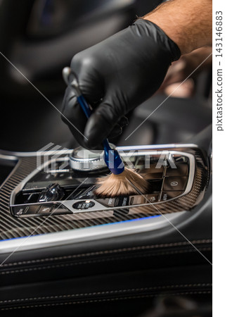 A gloved hand uses a soft detailing brush to clean a luxury car center console, showing carbon fiber trim, a rotary dial, and glossy buttons with moody lighting. A gloved hand uses a soft detailing brush to clean a luxury car center console, showing carbon fiber trim, a rotary dial, and glossy buttons with moody lighting. 143146883