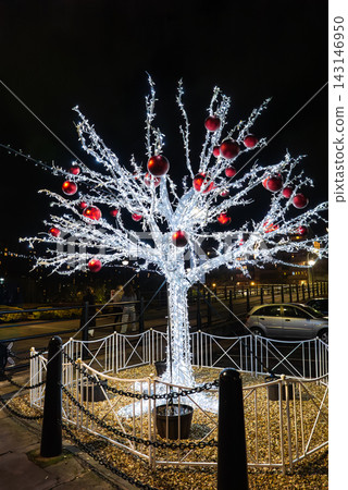 A white artificial ice tree with red baubles stands by the Royal Albert Dock in Liverpool at night, fenced in gravel, with pedestrians passing and warm lit buildings. A white artificial ice tree with red baubles stands by the Royal Albert Dock in Liverpool at night, fenced in gravel, with pedestrians passing and warm lit buildings. 143146950