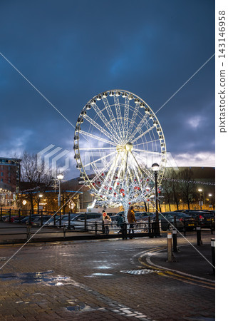Wheel of Liverpool near M and S Bank Arena rises over wet cobblestones as people gather by a Christmas tree with oversized red baubles during a festive winter evening. 143146958