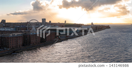 Aerial view at sunrise shows Liverpool, UK, the River Mersey, the Ferris wheel, ACC Liverpool and M and S Bank Arena, and Albert Dock warehouses under warm light and clouds. 143146986