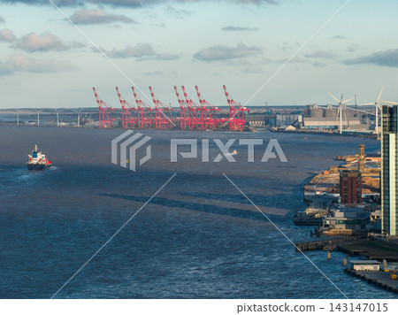 Aerial sunrise view in Liverpool shows River Mersey toward Port of Liverpool, bright red container cranes, docks, wind turbines, warehouses, ship and boat in morning light. 143147015
