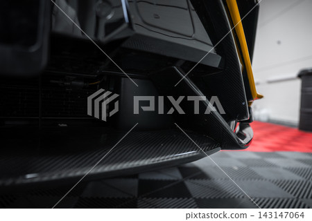 Close up of a yellow Lamborghini Urus Performante front aero, carbon fiber splitter and fin by the lower intake, indoors on a checkered garage floor with red matting. Close up of a yellow Lamborghini Urus Performante front aero, carbon fiber splitter and fin by the lower intake, indoors on a checkered garage floor with red matting. 143147064