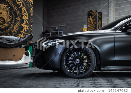 Close up three quarter view shows a black Mercedes Benz S Class with matte paint, gloss black wheels, and yellow calipers, indoors in a custom workshop with tools and wall art. Close up three quarter view shows a black Mercedes Benz S Class with matte paint, gloss black wheels, and yellow calipers, indoors in a custom workshop with tools and wall art. 143147082