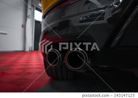 Close up of a yellow Lamborghini Urus Performante rear bumper shows dual round exhausts in a carbon fiber diffuser on a red and black checkered garage floor with moody lighting. Close up of a yellow Lamborghini Urus Performante rear bumper shows dual round exhausts in a carbon fiber diffuser on a red and black checkered garage floor with moody lighting. 143147125