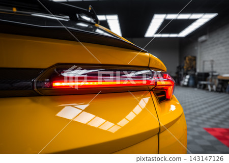 Close up shows the yellow Lamborghini Urus Performante rear left taillight glowing red under a black spoiler, with checkered floor, benches, and bright ceiling lights indoors. Close up shows the yellow Lamborghini Urus Performante rear left taillight glowing red under a black spoiler, with checkered floor, benches, and bright ceiling lights indoors. 143147126