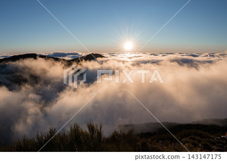 Sun rises above Pico do Arieiro on Madeira Island, Portugal, igniting mist over ridges. Cloud inversion spreads across slopes, shrubs frame the scene in crisp morning air. Sun rises above Pico do Arieiro on Madeira Island, Portugal, igniting mist over ridges. Cloud inversion spreads across slopes, shrubs frame the scene in crisp morning air. 143147175