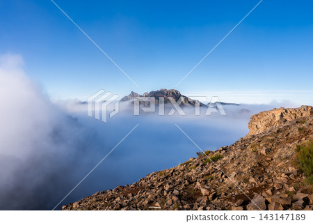 Jagged Pico do Arieiro rises above a sea of clouds in Madeira, Portugal, under warm morning light, with mist in valleys and wide angle view emphasizing elevation. Jagged Pico do Arieiro rises above a sea of clouds in Madeira, Portugal, under warm morning light, with mist in valleys and wide angle view emphasizing elevation. 143147189