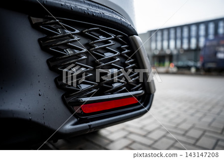 A close up shows the Lexus RX 450h plus rear bumper, glossy black lattice vent, and slim red reflector. Raindrops sit on dark paint near blurred buildings and parked cars. A close up shows the Lexus RX 450h plus rear bumper, glossy black lattice vent, and slim red reflector. Raindrops sit on dark paint near blurred buildings and parked cars. 143147208