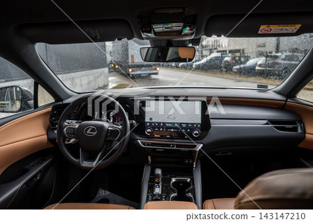 Driver view of Lexus RX 450h Plus cabin in urban setting, rainy day. Leather wheel, paddle shifters, wide nav display, glossy console, parked cars visible. Driver view of Lexus RX 450h Plus cabin in urban setting, rainy day. Leather wheel, paddle shifters, wide nav display, glossy console, parked cars visible. 143147210