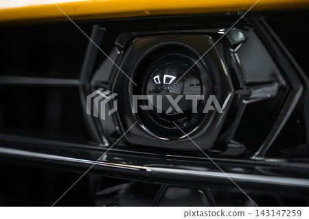 An extreme close up shows the front camera in a yellow Lamborghini Urus Performante grille, its glossy hexagonal housing reflecting ceiling lights in an indoor studio. An extreme close up shows the front camera in a yellow Lamborghini Urus Performante grille, its glossy hexagonal housing reflecting ceiling lights in an indoor studio. 143147259