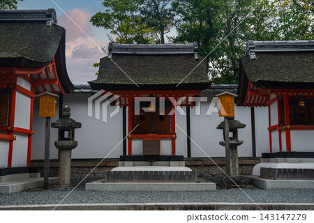 Small Shinto shrine buildings in Kyoto stand against a white wall, with stone lanterns and wooden placards. Soft evening light and pastel tones convey a calm, reverent mood. Small Shinto shrine buildings in Kyoto stand against a white wall, with stone lanterns and wooden placards. Soft evening light and pastel tones convey a calm, reverent mood. 143147279