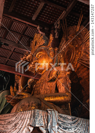 A gilded Buddhist statue glows in low interior light in Nara, Japan, with ornate crown, halo, layered robes, and hand mudras seen from a close upward angle. A gilded Buddhist statue glows in low interior light in Nara, Japan, with ornate crown, halo, layered robes, and hand mudras seen from a close upward angle. 143147287