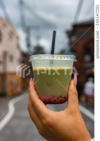 A hand holds a clear cup with iced matcha over red azuki and a smiley face, on a narrow Kyoto street with machiya and power lines in overcast light, shallow depth of field. A hand holds a clear cup with iced matcha over red azuki and a smiley face, on a narrow Kyoto street with machiya and power lines in overcast light, shallow depth of field. 143147291